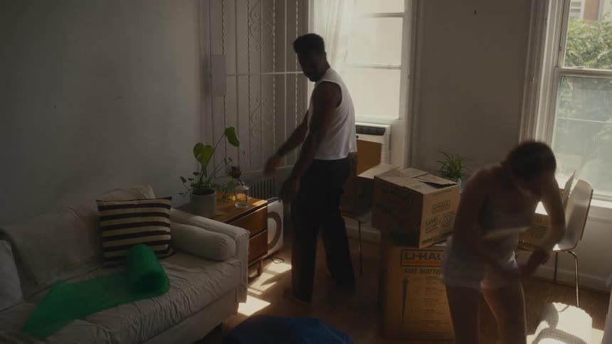 A man and a woman gather their personal belongings and pack them into cardboard boxes while preparing for a move in a bright apartment during the day