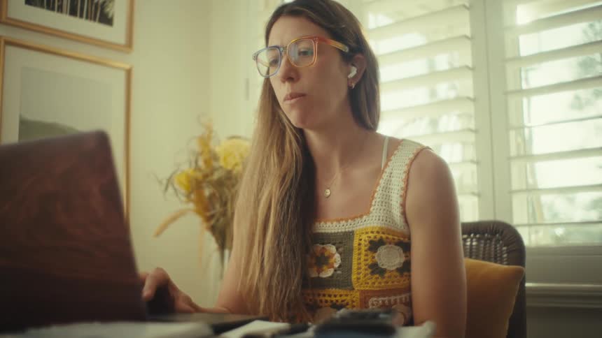 A woman wearing yellow glasses and a crochet top sits at a desk while speaking and gesturing toward her laptopthe room is brightly lit by natural light coming through window shutters and she smiles while appearing to be on a video call