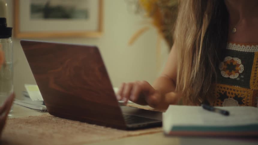 A woman wearing colorful glasses and a crochet top is shown alternating between studying a book and typing on her laptop the scene captures her focused academic work in a warm brightly lit indoor setting