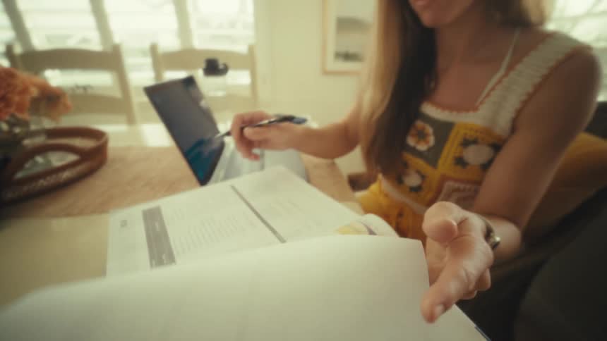 A woman in a yellow patterned dress sits at a sunlit table holding a pen and turning a page of an open book with a laptop beside her
