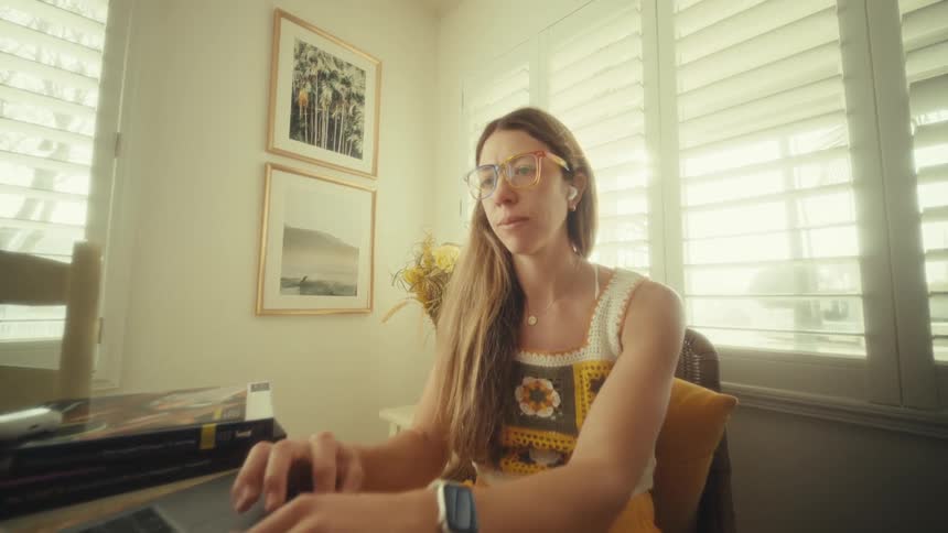 A woman is sitting at a desk in a sunlit room typing on a laptop as she reads the screen