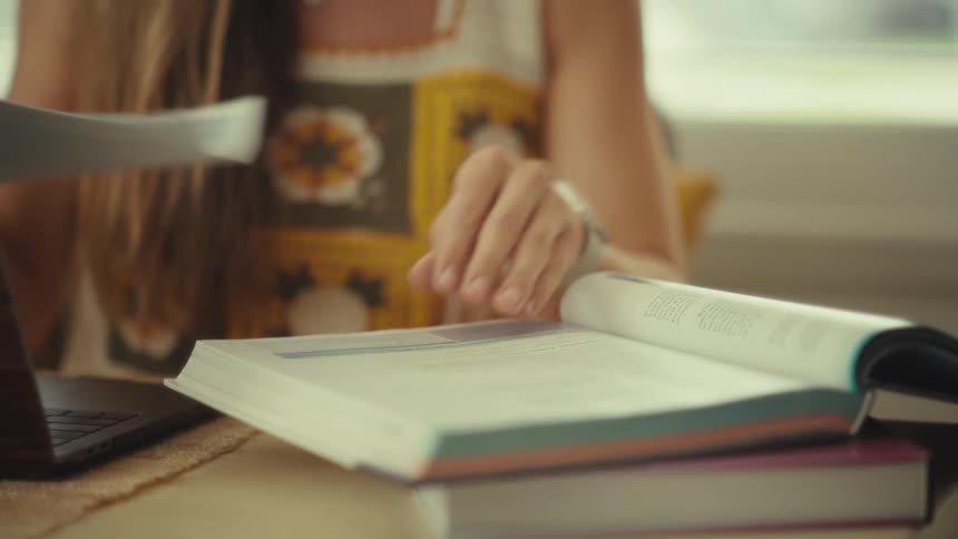 A young woman with glasses sits at a wooden desk and focuses on her work by typing on a laptop and turning pages of a large textbook to take notes with a black pen in a bright room