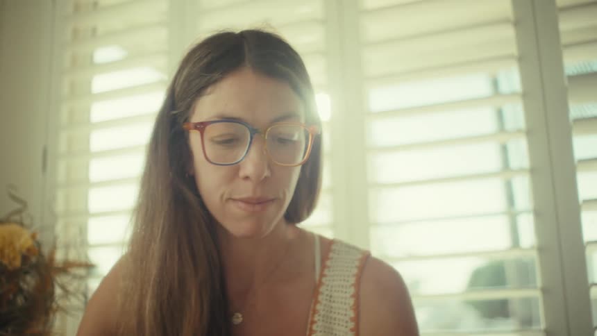 A woman with long brown hair and colorful glasses looks down thoughtfully as sunlight filters through white window shutters behind her