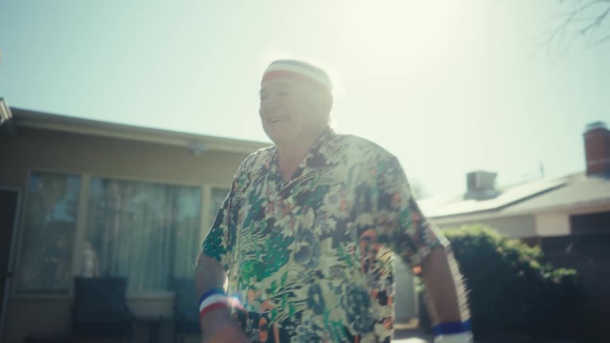 An elderly man wearing a floral shirt and patriotic wristbands moves energetically in a sunny backyard the bright sunlight creates a lens flare as he smiles and appears to be dancing or exercising outdoors