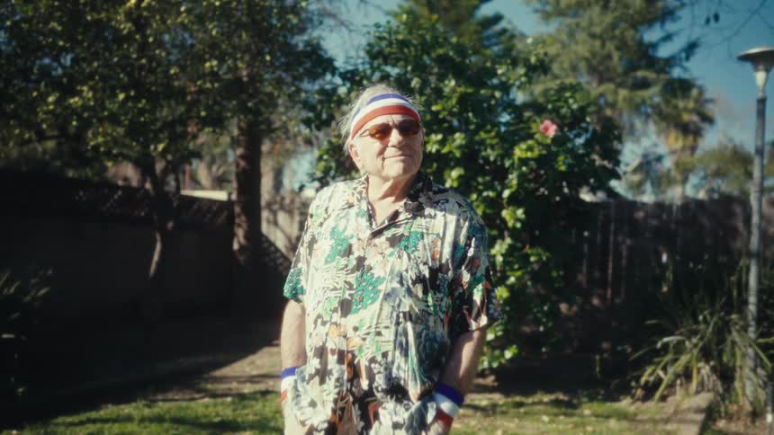 Elderly man smiles as he stands in his yard with his hands in his shorts pockets