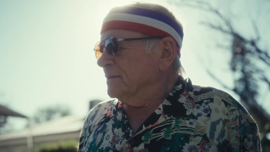 An elderly man wearing a floral shirt and headband stands outdoors partially silhouetted by bright sunlight