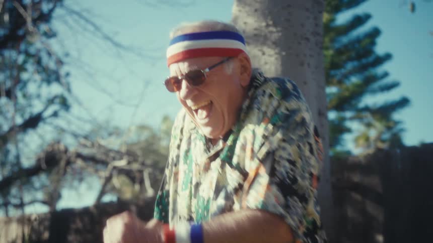 Elderly man wearing a sports headband uses a leaf blower in the backyard beside a wooden fence