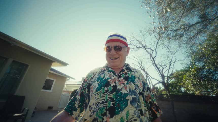 An elderly man holding a leaf blower smiling as he moves the tool around the backyard lawn on a sunny day