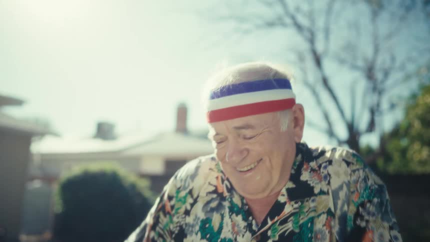 Elderly man wearing a sports headband dances in the backyard under sunlight