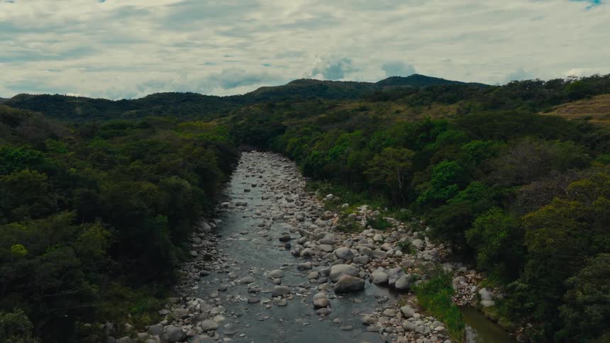 An aerial view shows a shallow rocky river flowing through a lush green valley under a cloudy sky distant mountains rise behind the dense forest that lines both sides of the riverbed
