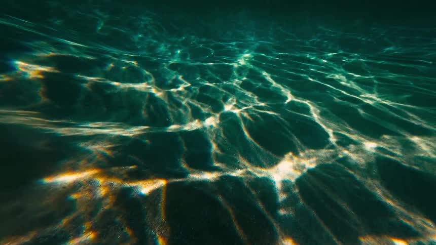 Luminous sunlight penetrates the teal water to cast vibrant dancing caustic patterns across a golden sandy seabed which eventually gives way to the deep silent shadows of the open ocean