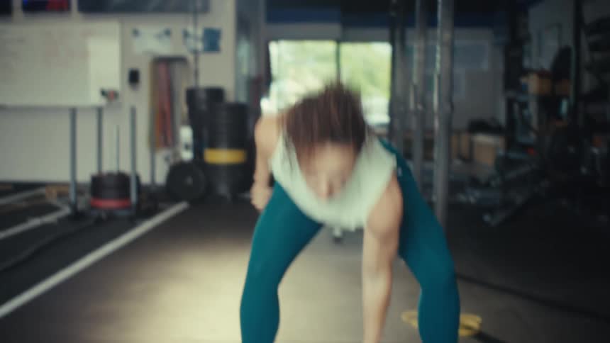 A hispanic woman with curly hair performs a kettlebell snatch inside a gym and she wears a white top and blue leggings while lifting the weight overhead with one arm