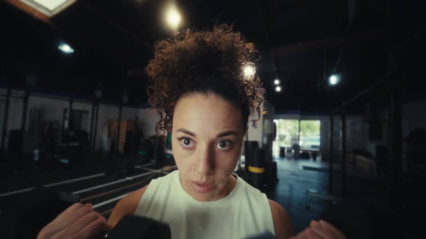 A woman with curly hair performs bicep curls using black dumbbells inside a gym