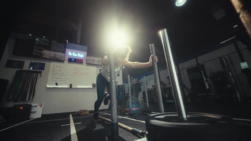 A woman with curly hair pushes a weighted sled across a gym floor while other athletes train in the background the shot starts with a closeup of her face and pulls back to show her fullbody effort against the resistance