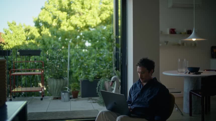A man wearing a dark blue shirt and beige pants sits in a chair indoors while working on a laptop with a view of a balcony and green trees in the background