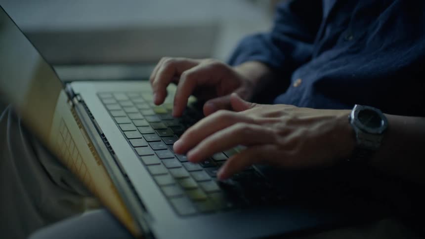 In a dimly lit setting a persons hands are shown typing steadily on the keyboard of a dark laptopthe individual is dressed in a blue buttondown shirt and lightcolored trousers wearing a silver watch on their left wrist