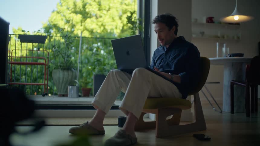 Man sitting on a chair uses a laptop near a glass door with a view of plants