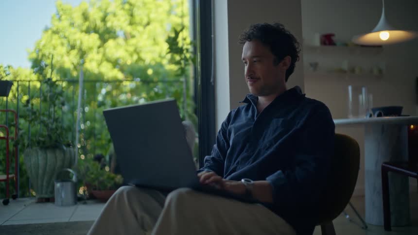 A young man sitting on a stool near his corridor typing on his laptop at midday with trees and green vegetation visible outside the building