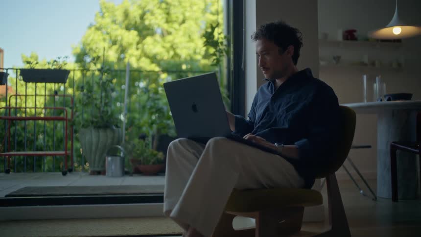 A man with dark hair sits in a chair and works on a laptop while drinking from a black mug inside a room next to an open door that leads to a balcony with green plants