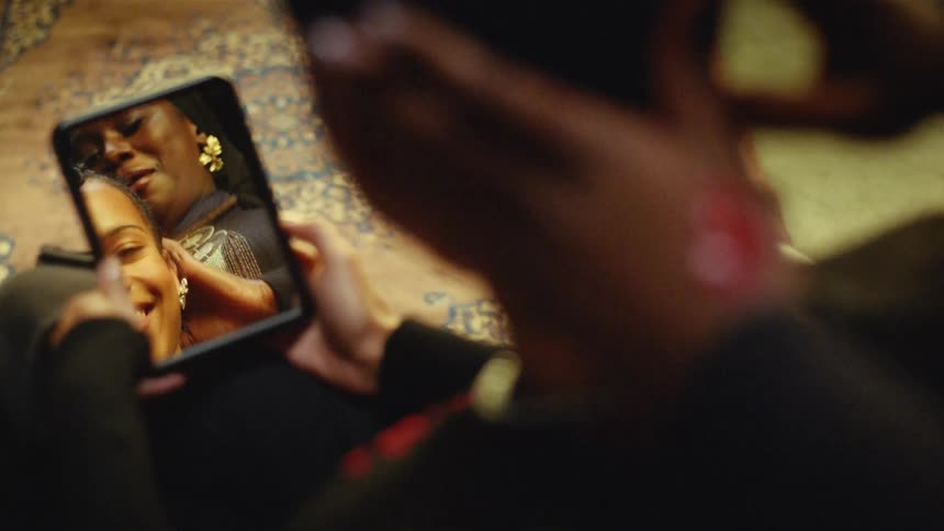 A view from behind a woman shows her reflection in a small hand mirror as a friend adjusts her hair while they sit together in a room with patterned rugs