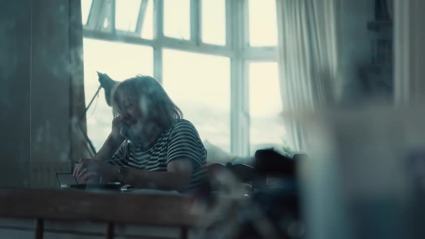 An older woman with long hair wears a striped shirt while sitting at a desk talks on a phone and writes in a notebook with large windows and a lamp in the background