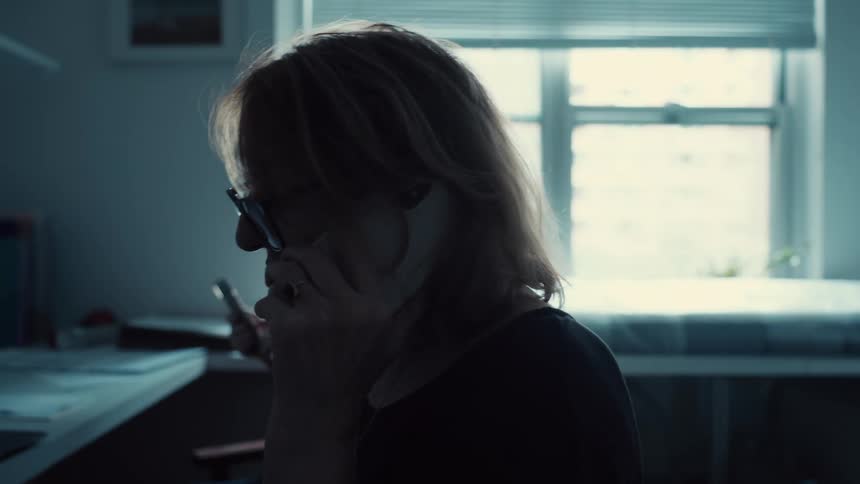 A side view of a woman with glasses in a dark room shows her talking on a phone with a blurred window and a medical bed in the background