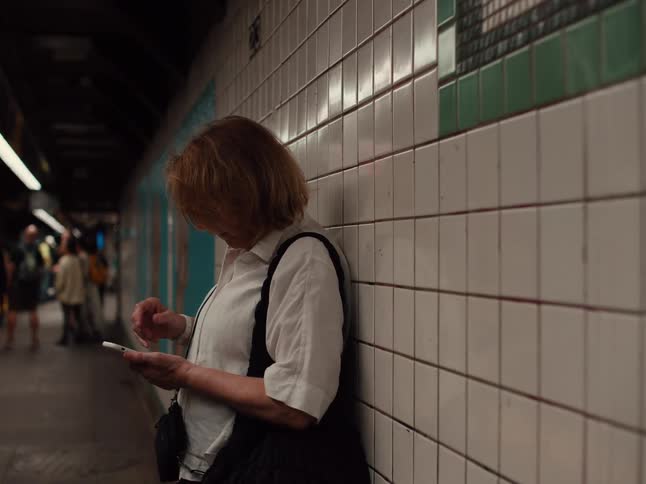 Person leaning against a tiled wall in a subway station while using a cellphone