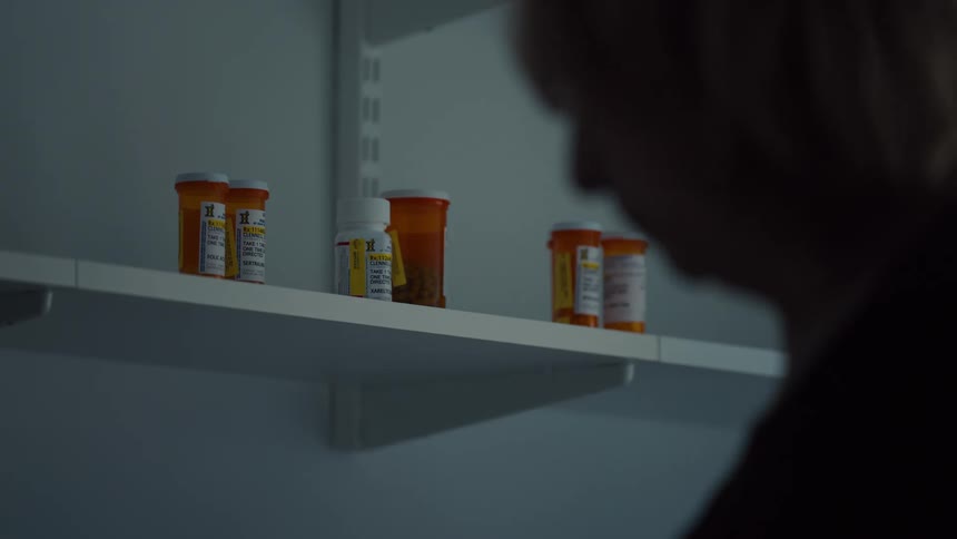 An elderly woman standing by a counter with several pill containers on it drinking from a coffee mug