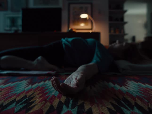 A woman lies flat on her back on a patterned rug in a dimly lit room her eyes closed and arms resting at her sides in a relaxed position a single desk lamp glows in the background casting a soft light over the quiet domestic scene