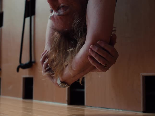A close view shows a person hanging upside down in a fitness studio with their hands clasped behind their head while wearing a watch and a ring against a light wood wall and floor