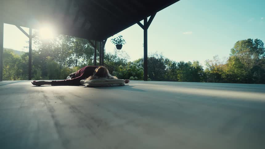 Adult woman wearing a red sweater doing yoga on a porch surrounded by trees outdoors during day time