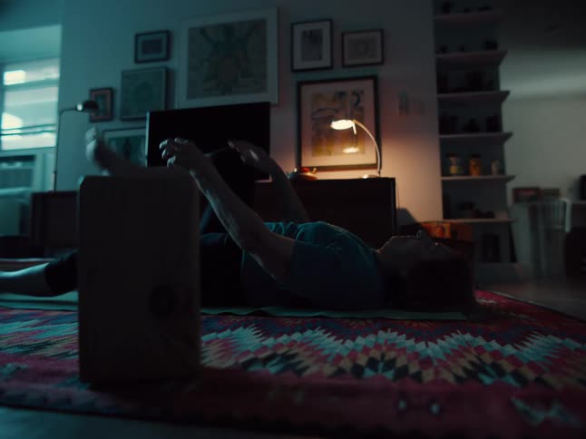 Woman performs stretching while lying on a rug in a room lit by a lamp