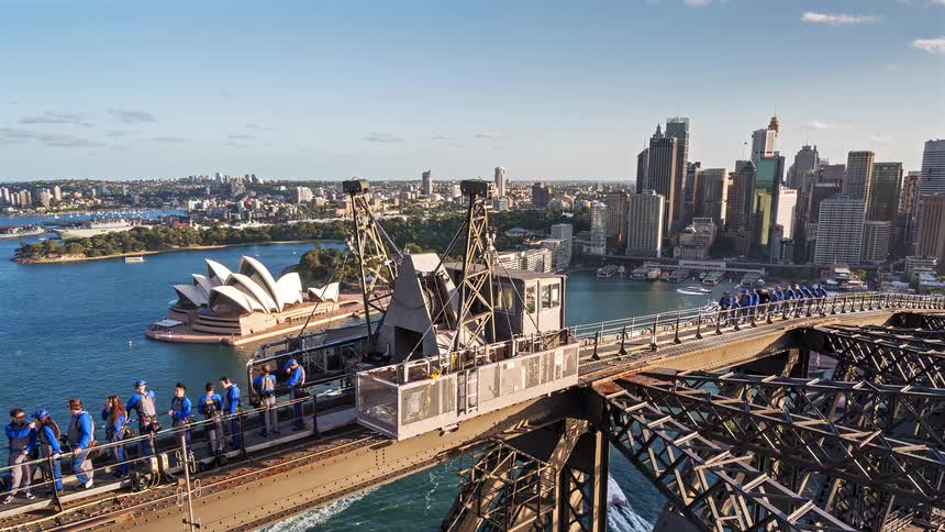 A high angle view shows people walking across the steel structure of the sydney harbour bridge with the sydney opera house and city skyline in the background