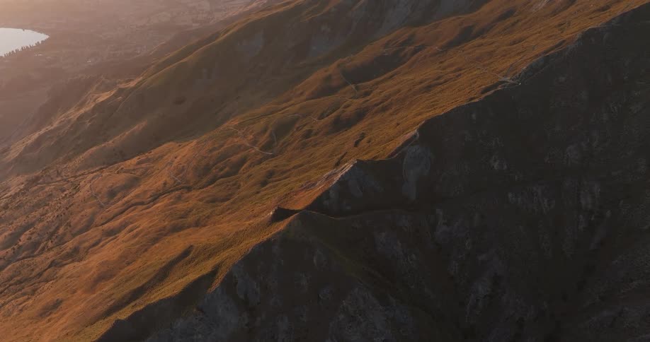 An aerial view shows the golden slopes of a mountain ridge during a sunset with a small person walking along a narrow trail at the top