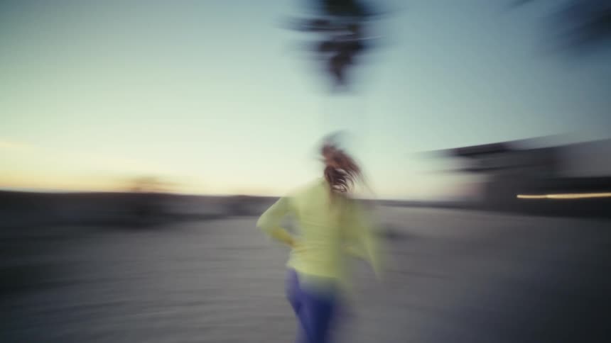 A woman wearing a yellow jacket and blue leggings runs away from the camera along a concrete beach path at dusk