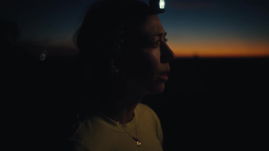 A side view shows a pregnant woman wearing a headlamp and a light shirt standing in a dark field while looking toward a sunset on the horizon