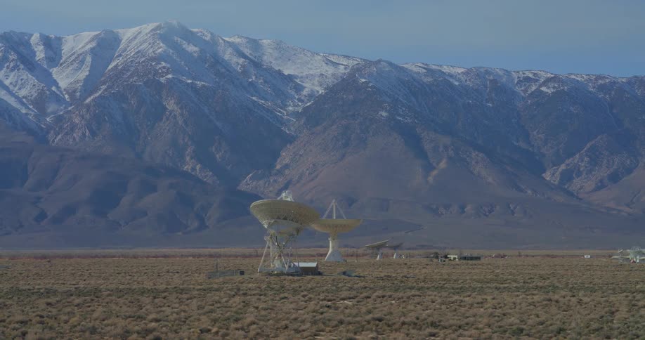 Large white radio telescopes stand in a vast desert valley while the camera pans across the arid terrain toward high mountains covered in snow under a clear sky at a remote scientific research facility