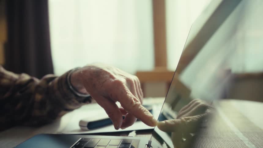 Elderly man uses a laptop while handling papers on a table indoors