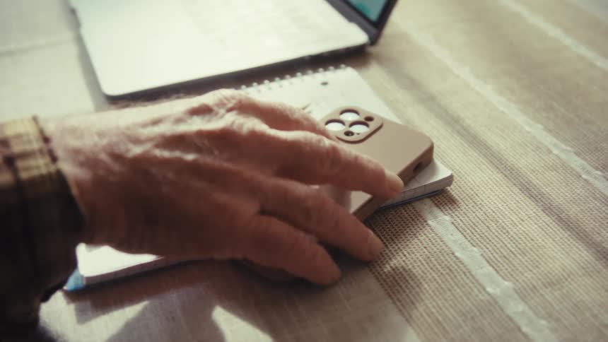 Elderly man sitting at a table facing a laptop picks up his smartphone