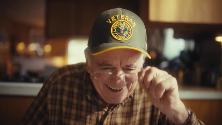 An elderly man wearing a veteran cap and glasses smiles warmly as he looks down at something on the table in front of him then removes his eyeglasses