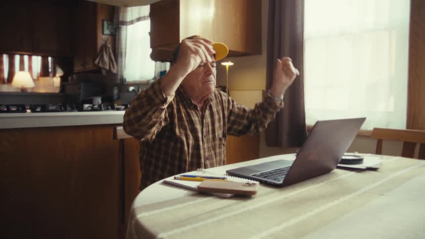 An older man wearing a plaid shirt and a baseball cap yells and raises his arms while sitting at a table with a laptop computer inside a home