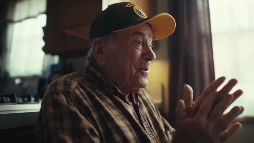 An elderly man wearing a plaid shirt and a yellow brimmed hat sits in a kitchen while laughing and clasping hands