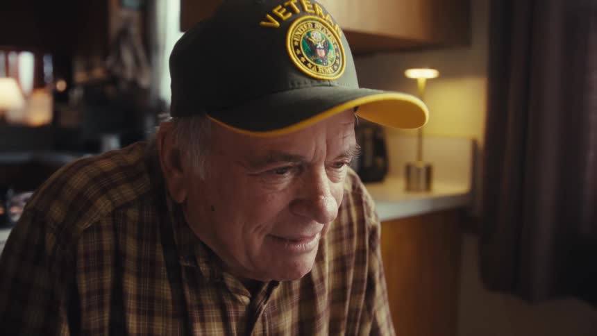 Elderly man wears a cap with the word veteran while sitting indoors