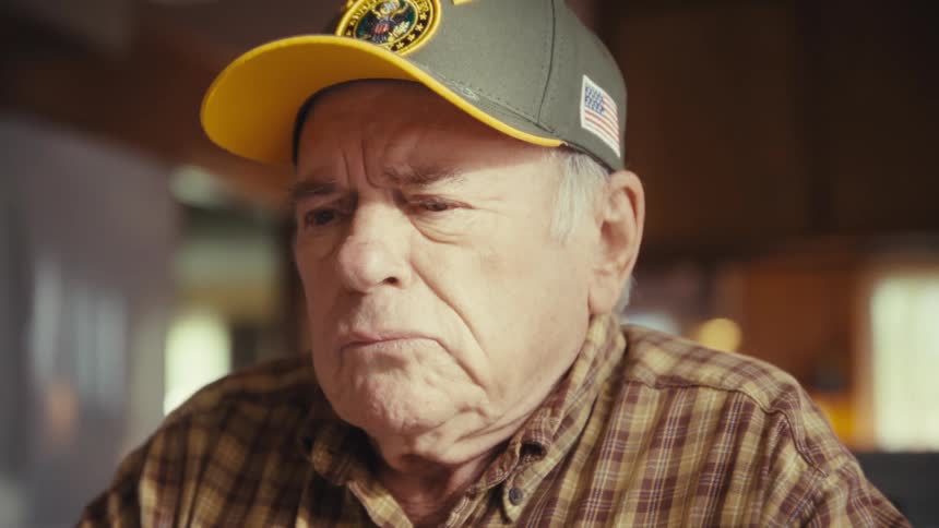 An elderly man wearing a veteran baseball cap and a plaid shirt sits in a kitchen while looking intently at a laptop screen with a serious expression as blurred wooden cabinets and a white refrigerator stand in the background
