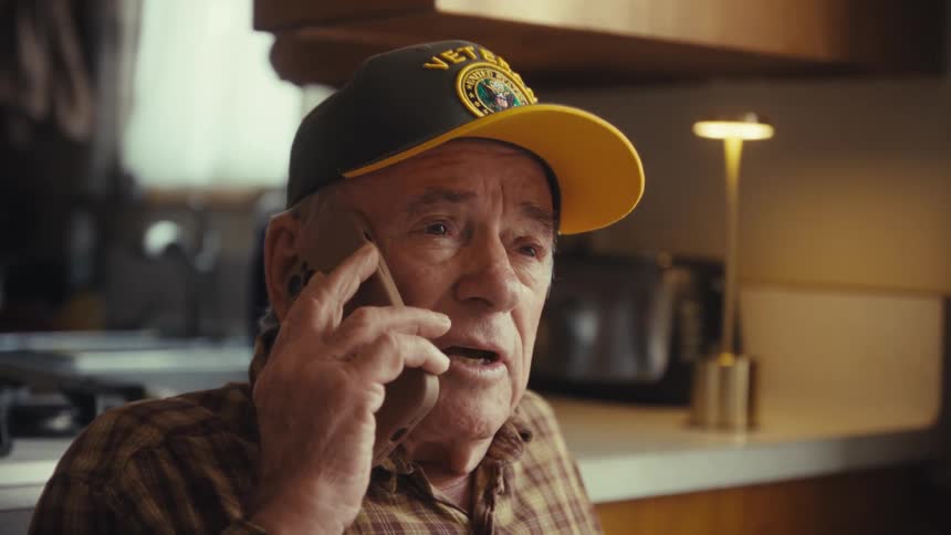 An older man wearing a plaid shirt and a baseball cap holds a mobile phone to his ear while talking and looking toward the side inside a kitchen