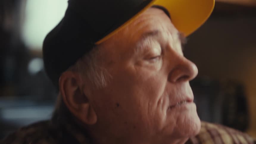 An elderly man wearing a baseball hat sitting at a dining table dancing and shaking his body having fun with a laptop in front of him at midday
