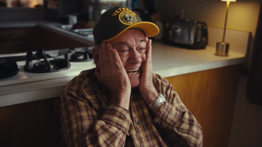 An older man wearing a plaid shirt and a baseball cap laughs with his hands on his face while sitting in a kitchen