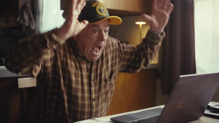An older man wearing a plaid shirt and a baseball cap yells and raises his arms while sitting at a table with a laptop computer inside a home