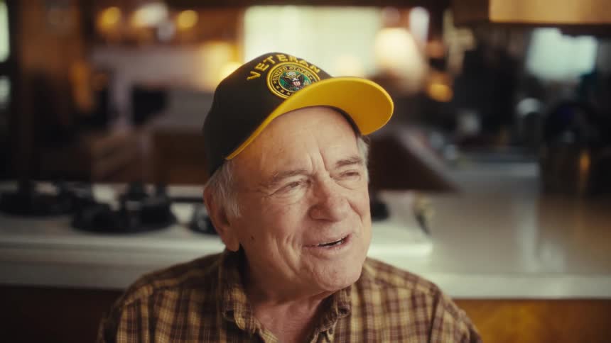 An elderly man sitting at a dining table at midday smiling as he takes off his glasses with a laptop in front of him and a hat that says veteran on his head