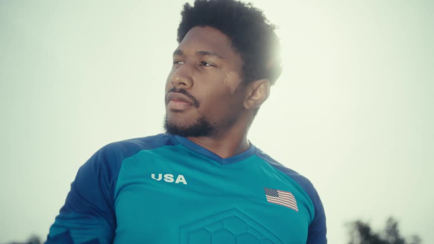 A male athlete wearing a blue jersey standing in an open field on a sunny day